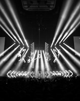 A black and white image captures a dramatic indoor scene featuring a large stage setup with intense beams of light radiating from the center. Two large screens are prominently displaying the text '01:33 ESL One Cologne 2017'. The audience can be seen seated in the dark foreground, while angular patterns can be spotted on the stage.